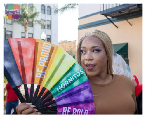 A person holding a colorful fan with the inscriptions "be proud" and "be bold" at an outdoor event.