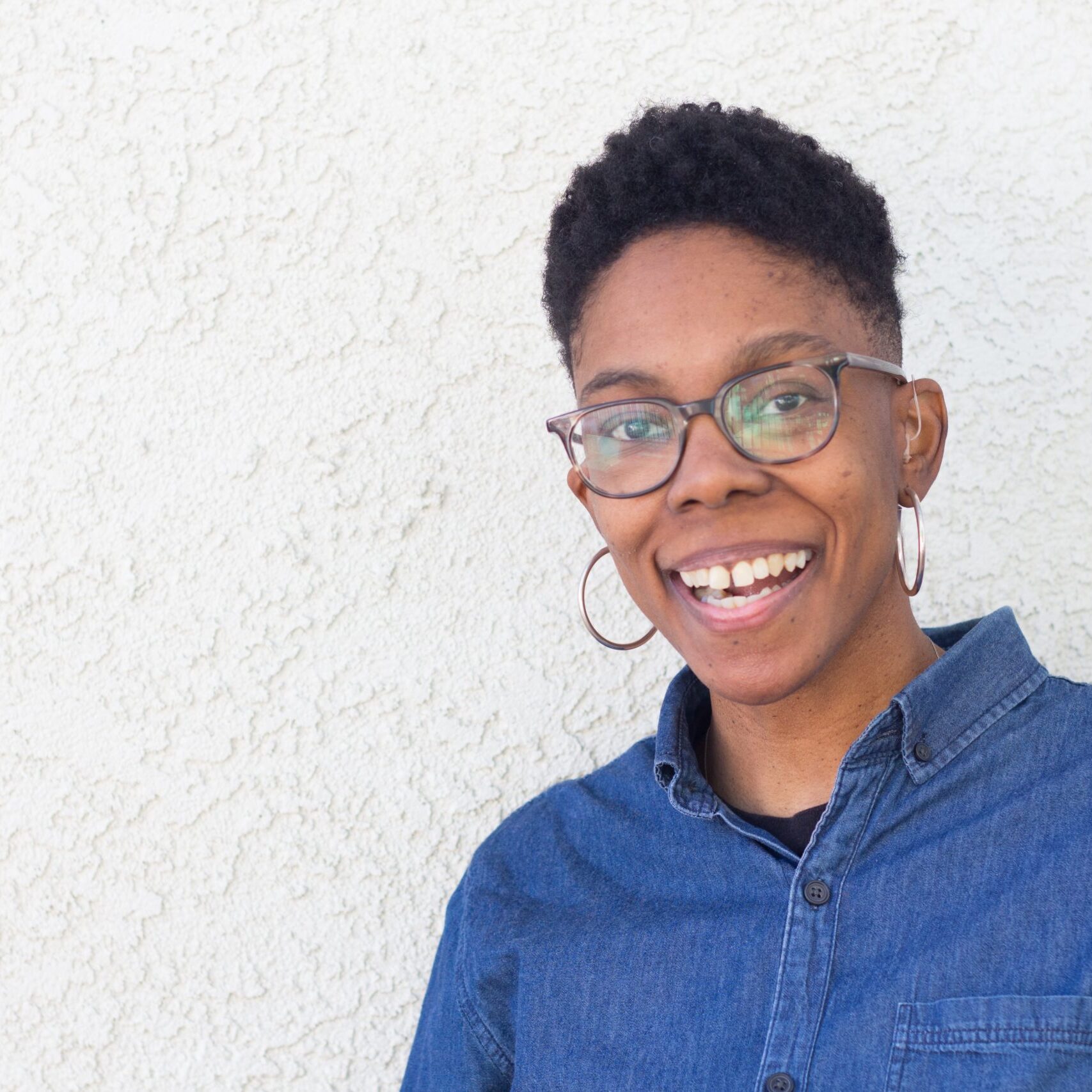 A smiling person with short hair and glasses, wearing a denim shirt and hoop earrings, stands against a textured white wall.