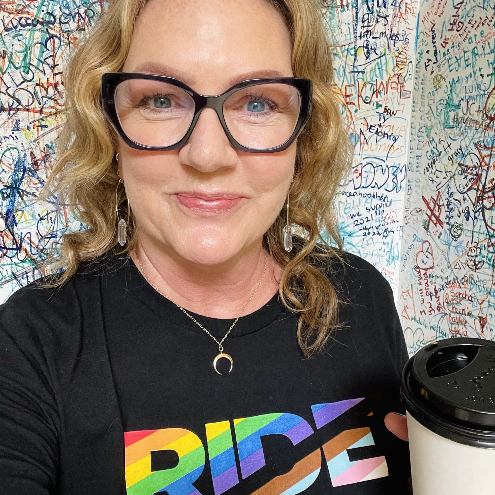 Woman with glasses smiling, holding a coffee cup, wearing a "ride" t-shirt, standing in front of a graffiti-covered wall.