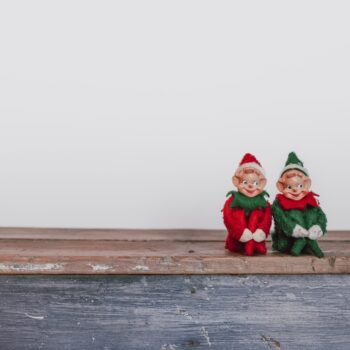 Two christmas elf decorations sitting on a wooden surface against a white background.